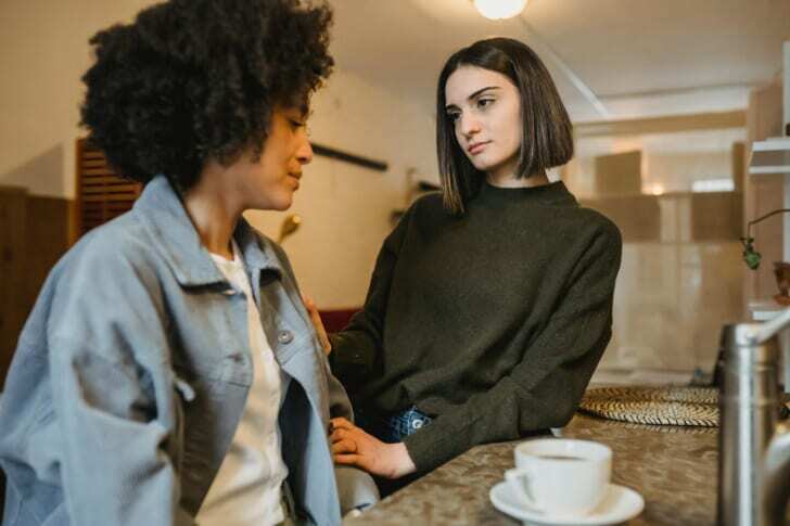 Delicate female holding hand of upset African American female having problem sitting at counter with cup of coffee